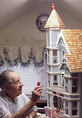 Eldery man painting and constructing a light blue Victorian dollhouse in a white room with Victorian style curtains