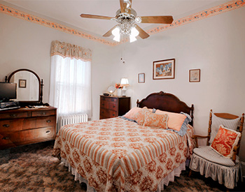 Bedroom with one bed with peach and white cover and a brown dresser and mirror set and one small chair with a light blue cushion