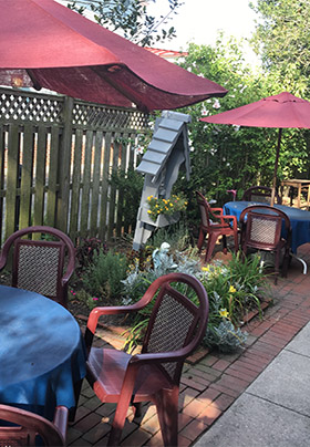 Outdoor dining set with red umbrellas and chairs and blue tables. Flowers surround the area and there is red brick and sidewalk on the ground.